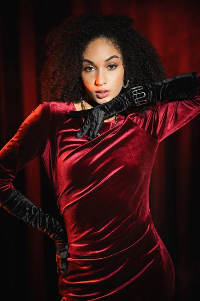 Mujer afroamericana de moda en vestido de terciopelo y guantes posando cerca de cortinas rojas - foto de stock