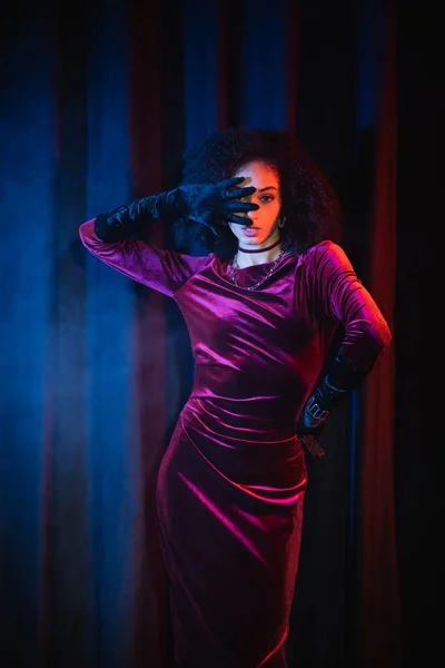 Modèle afro-américain tendance en robe de velours et gants sur fond rouge avec lumière bleue — Photo de stock