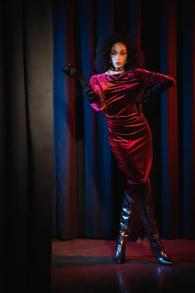 Full length of stylish african american woman in dress and knee boots posing on red background with blue light — Stock Photo