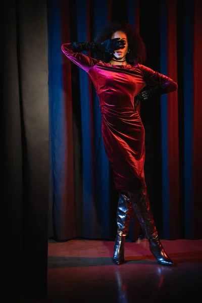 Volle Länge der trendigen afrikanisch-amerikanischen Frau im Kleid bedeckt Gesicht auf rotem Hintergrund mit blauem Licht — Stockfoto