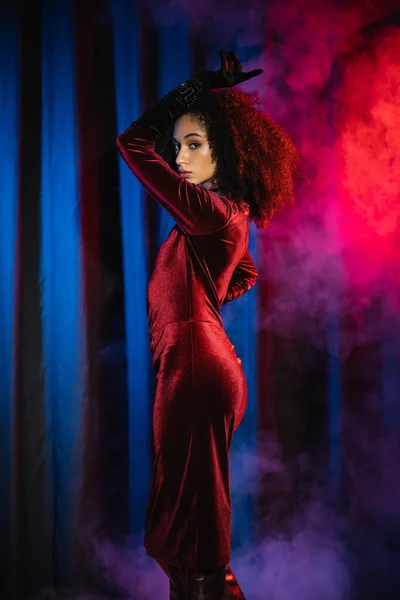 Femme afro-américaine élégante en robe de velours regardant la caméra sur fond de lumière bleue et de fumée — Photo de stock