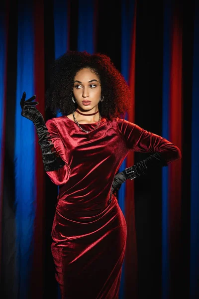 Donna afroamericana alla moda in guanti di velluto e vestito guardando altrove su sfondo rosso con luce blu — Foto stock