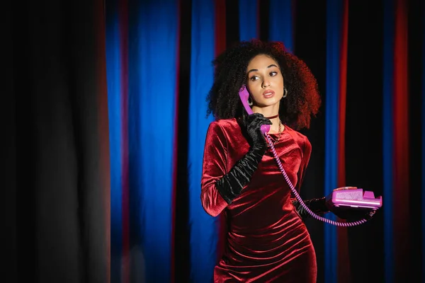 Élégante femme afro-américaine en robe de velours parlant sur téléphone rétro sur fond bleu avec de la lumière — Photo de stock