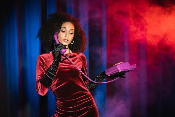 Mujer americana africana rizada en vestido hablando por teléfono sobre fondo rojo con luz azul y humo - foto de stock