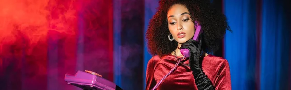 Elegante mujer afroamericana en guante de terciopelo hablando por teléfono sobre fondo rojo con luz azul con humo, pancarta - foto de stock