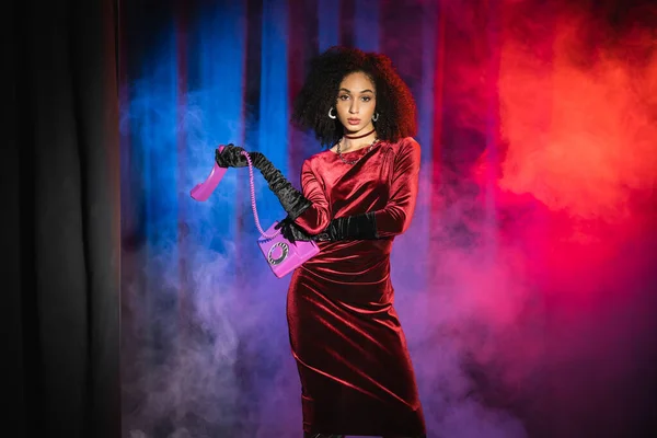 Joven modelo afroamericano en vestido de terciopelo y guantes con teléfono vintage cerca del fondo con luz azul y humo - foto de stock