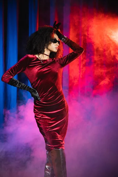 Модная африканская модель в бархатном платье и солнцезащитных очках позирует на красном фоне с голубым светом и дымом — стоковое фото