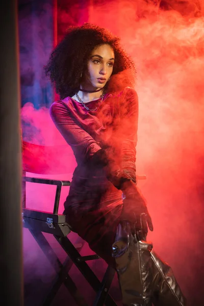 Trendy african american woman in velvet dress sitting on chair on red background with blue light and smoke — Stock Photo