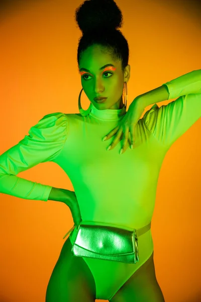 Trendy african american woman in neon bodysuit and makeup posing on orange background — Stock Photo
