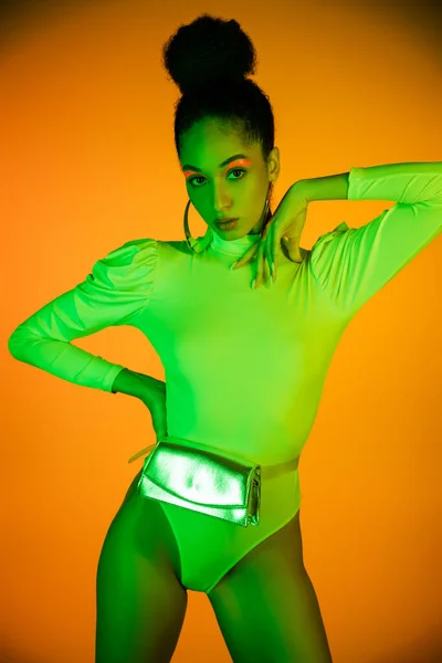 Stylish african american model with neon makeup and bodysuit standing on orange background — Stock Photo