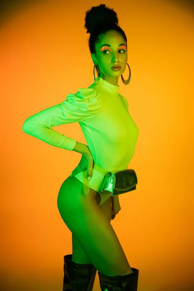Sexy african american woman in neon bodysuit and knee boots posing on orange background — Stock Photo