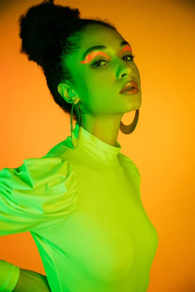 Ritratto di elegante donna afroamericana con eyeliner al neon che guarda la fotocamera su sfondo arancione — Foto stock