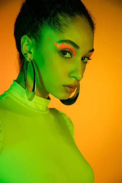 Modèle afro-américain à la mode avec eye-liner néon regardant la caméra sur fond orange — Photo de stock