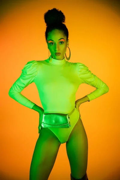 Trendy african american model with neon eyeliner and bodysuit holding hands on hips on orange background — Stock Photo