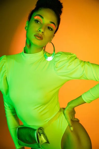 Fashionable african american model with neon visage posing in bodysuit on orange background — Stock Photo