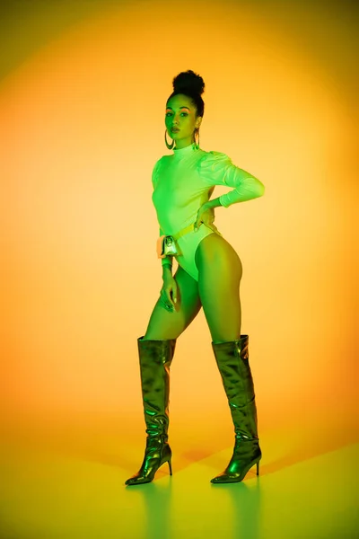 Full length of trendy african american model in neon bodysuit and knee boots on orange background — Stock Photo