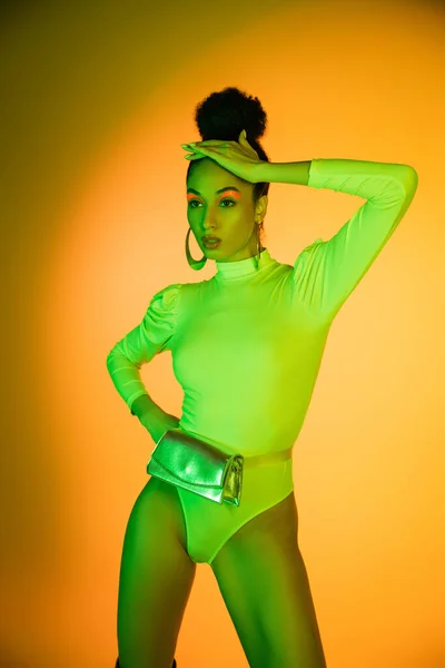 Stylish african american model in neon bodysuit touching hair on orange background — Stock Photo