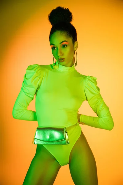 Pretty african american model in neon bodysuit and waist bag looking away on orange background — Stock Photo