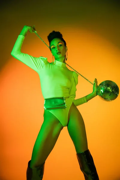 Fashionable african american woman in neon bodysuit and knee boots holding disco ball on orange background — Stock Photo