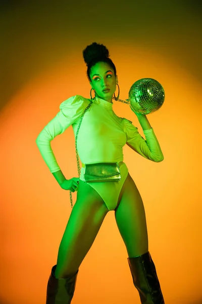 Trendy african american woman in neon bodysuit holding disco ball on orange background — Stock Photo