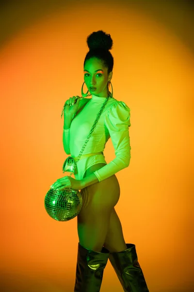 Модная африканская американка в неоновом костюме и ботинках на коленях держит диско-мяч на оранжевом фоне — стоковое фото