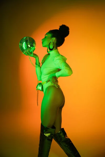 Вид сбоку на сексуальную афроамериканку в неоновом костюме, смотрящую на дискотечный мяч на оранжевом фоне — стоковое фото