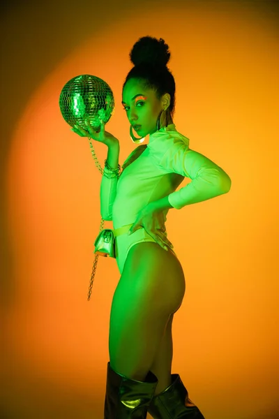 Модная африканская модель в неоновом костюме и ботинках на коленях, держащая диско-мяч и смотрящая в камеру на оранжевом фоне — стоковое фото