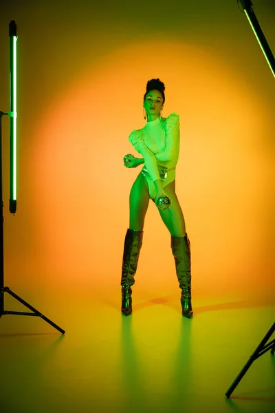 Stilvolle afrikanisch-amerikanische Frau in neonfarbenem Body und Kniestiefeln, die Discokugeln neben Leuchtstofflampen auf orangefarbenem Hintergrund hält — Stockfoto