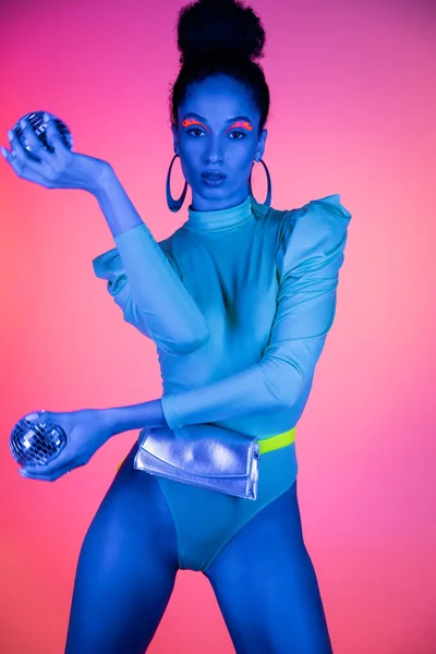 Modèle afro-américain à la mode avec maquillage au néon et body tenant des boules disco sur fond rose — Photo de stock