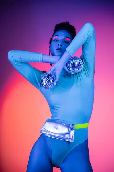 Модная африканская модель с неоновым глазком позирует с диско-шариками на розовом и фиолетовом фоне — стоковое фото