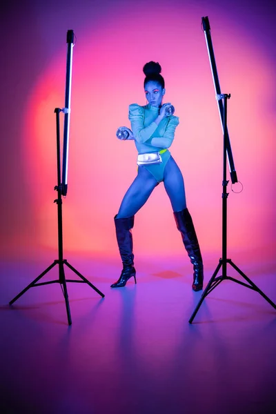 Trendy african american model in bodysuit holding disco balls near neon lamps on purple background — Stock Photo