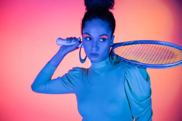 Дивна американка з неоновими очима тримає тенісну ракетку на рожевому фоні. — стокове фото