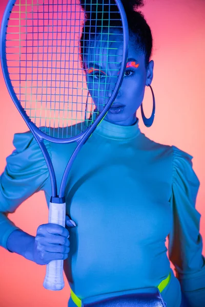 Portrait of fashionable african american woman with neon eyeliner holding tennis racket on pink background — Stock Photo