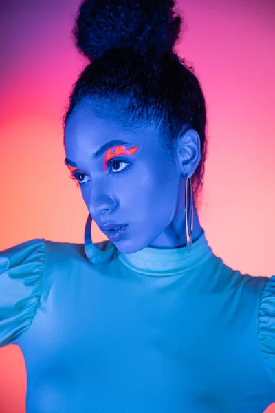 Ritratto di modello afroamericano alla moda con eyeliner al neon che distoglie lo sguardo su sfondo rosa e viola — Foto stock