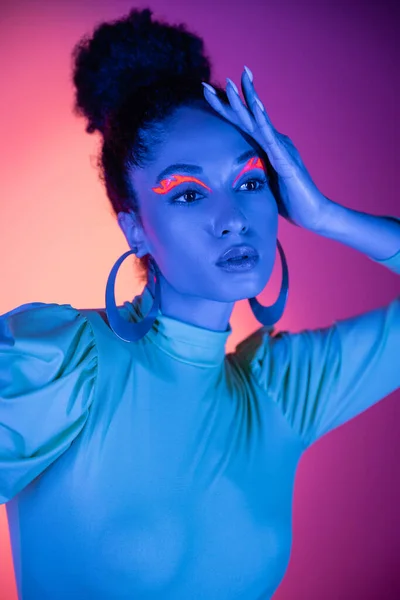 Portrait of fashionable african american model with neon makeup touching hair on pink and purple background — Stock Photo