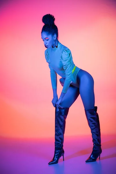 Longitud completa de la mujer afroamericana de moda con maquillaje de neón y body ajustando la bota de rodilla sobre fondo rosa y púrpura - foto de stock