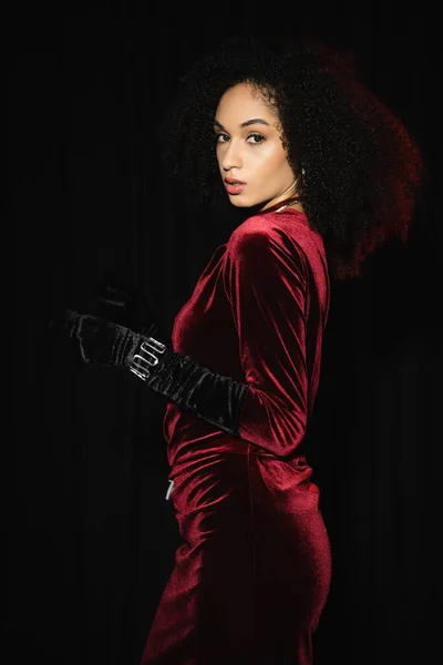 Trendy african american woman in velvet burgundy dress and glove looking at camera isolated on black — Stock Photo