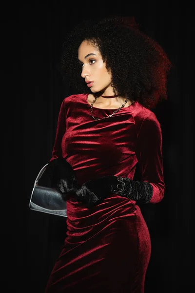 Mulher americana africana na moda em vestido de borgonha e luvas de veludo segurando bolsa isolada em preto — Fotografia de Stock