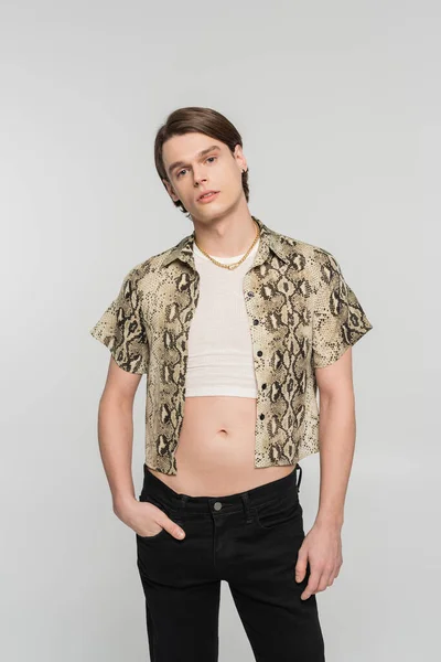 Stylish bigender person in snakeskin print blouse posing with hand in pocket of black pants isolated on grey — Stock Photo