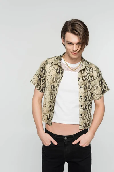 Young and trendy bigender model in animal print blouse posing with hands in pockets of black pants isolated on grey — Stock Photo