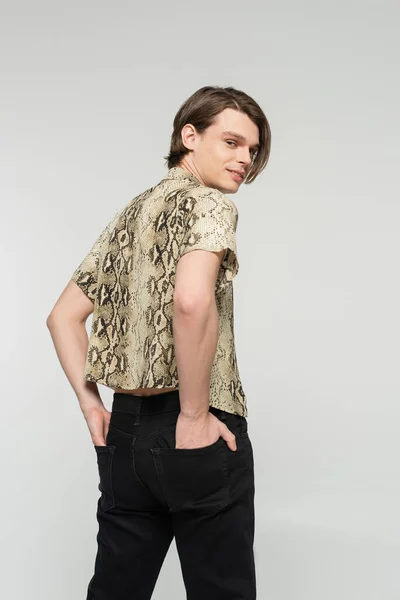 Trendy pangender person in animal print blouse holding hands in back pockets and smiling at camera isolated on grey — Stock Photo