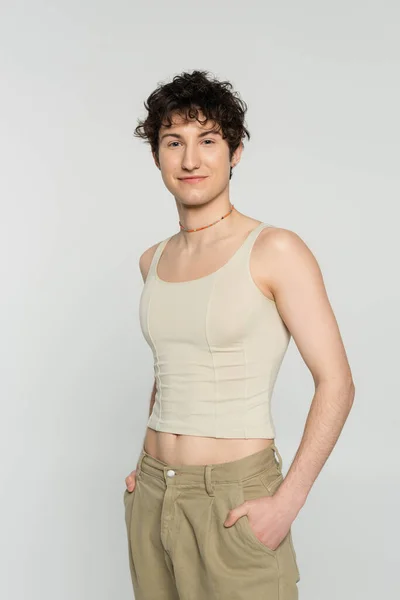 Brunette bigender person in tank top standing with hands in pockets of beige pants and smiling at camera isolated on grey — Stock Photo