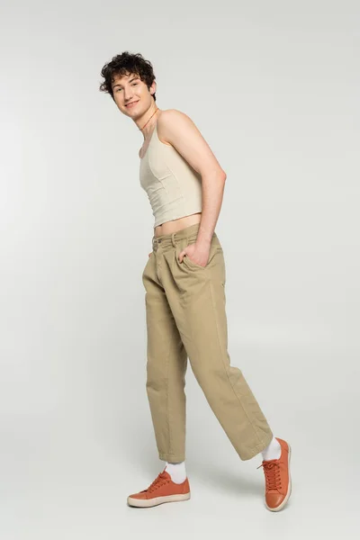 Persona spensierata bigender in canotta e sneakers in posa con mano in tasca di pantaloni beige su sfondo grigio — Foto stock