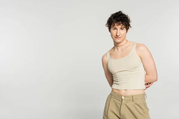 Young bigender model in tank top and pants standing with hands behind back isolated on grey — Stock Photo