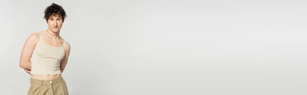 Brunette modèle pansexuel en débardeur posant avec les mains derrière le dos isolé sur gris, bannière — Photo de stock