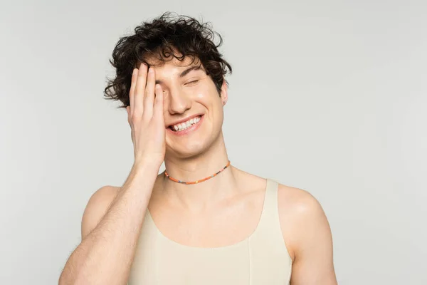 Cheerful nonbinary model in tank top and beads covering face with hand and smiling with closed eyes isolated on grey — Stock Photo