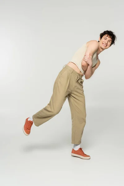 Pleine longueur de personne gaie non binaire en pantalon beige et baskets posant avec les bras croisés sur fond gris — Photo de stock