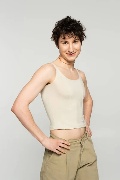 Trendiges und glückliches Pangender-Model, das mit den Händen auf den Hüften steht und isoliert auf grau in die Kamera lächelt — Stockfoto
