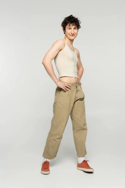 In voller Länge glücklich bigender Modell in beige Hose posiert mit Händen auf Hüften auf grauem Hintergrund — Stockfoto