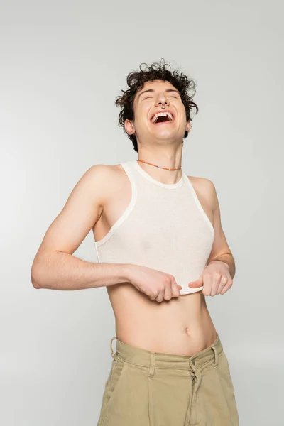 Excited bigender person pulling down crop top and laughing with closed eyes isolated on grey — Stock Photo